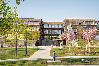 Vue extérieure avec une façade en bois, une aire de jeux, des espaces verts et des escaliers menant aux terrasses du campus éducatif à Vienne.